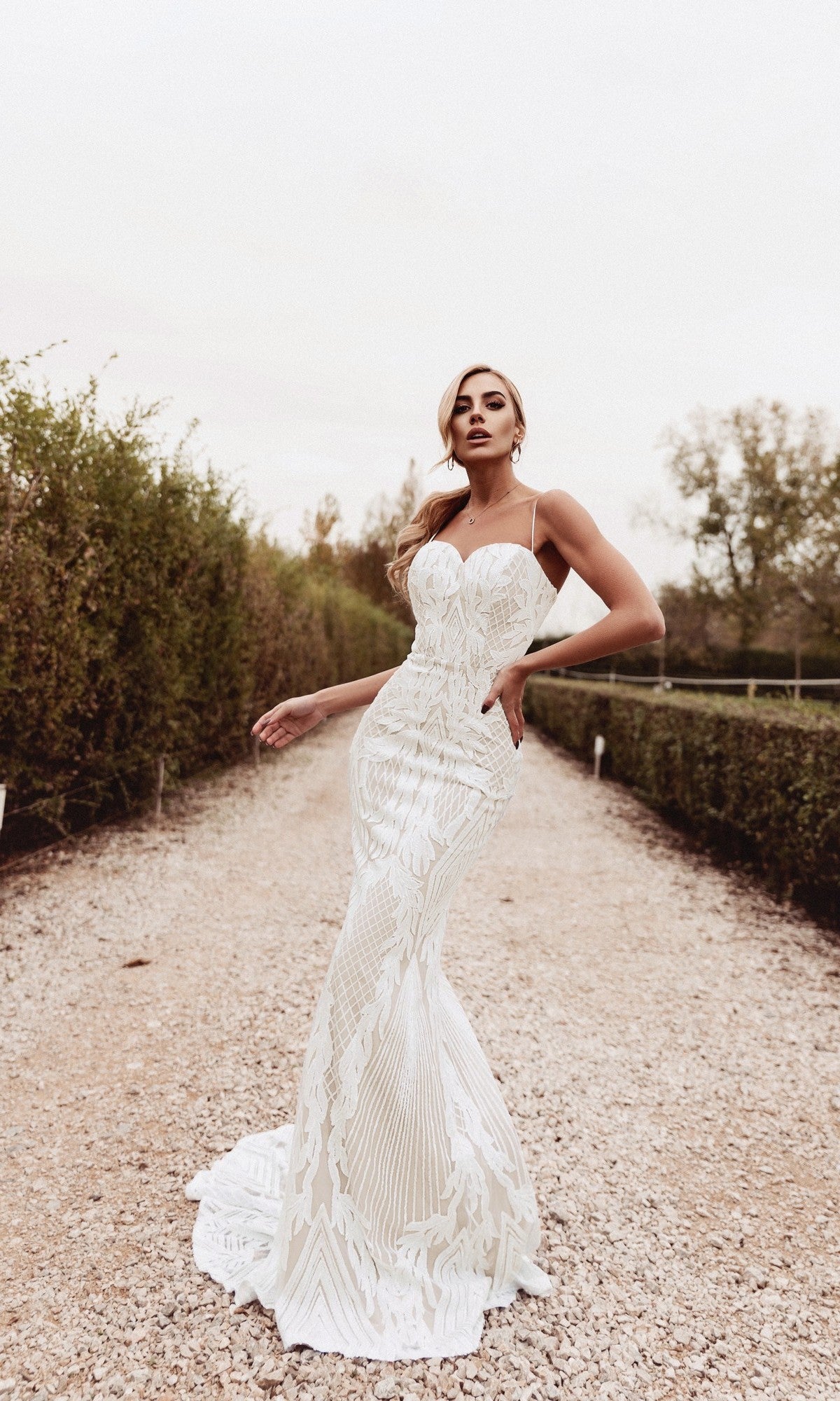 A woman stands outdoors on a gravel path wearing the Tina Holly BB050, a long white mermaid wedding dress featuring intricate patterns, a flared hem, and a form-fitting sweetheart neckline.